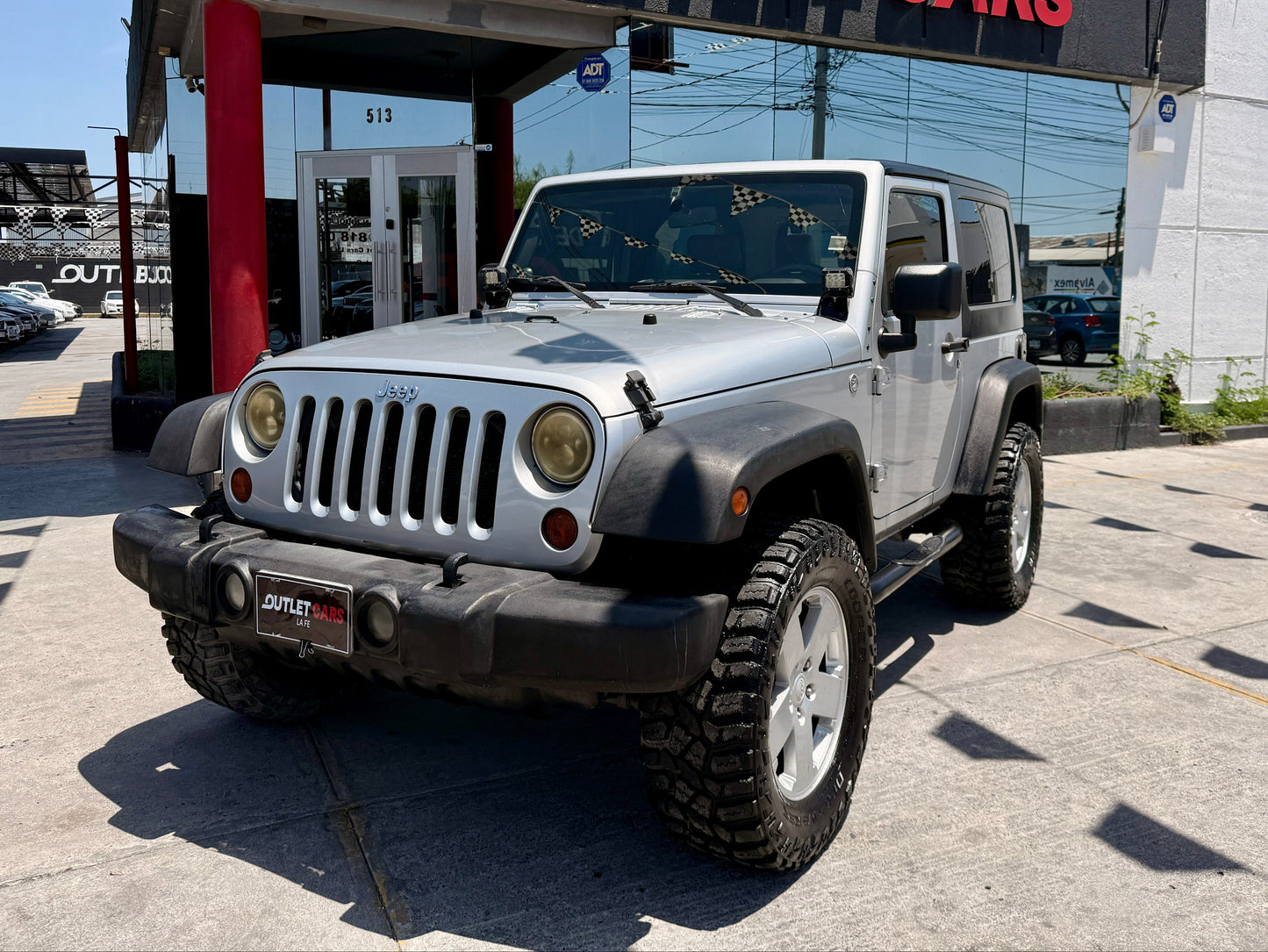 A silver Jeep Wrangler parked outdoors in front of an Outlet Cars dealership building on a paved lot.